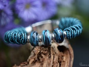 Heren gedenkarmband met turquoise-zwart geknoopte band en drie petrol-zwart glasbedels waarvan de middelste crematie-as bevat.