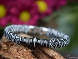 Heren gedenkarmband van zwart-wit paracord met zwarte design bedels waarvan de middelste rvs bedel crematie-as zichtbaar bevat.